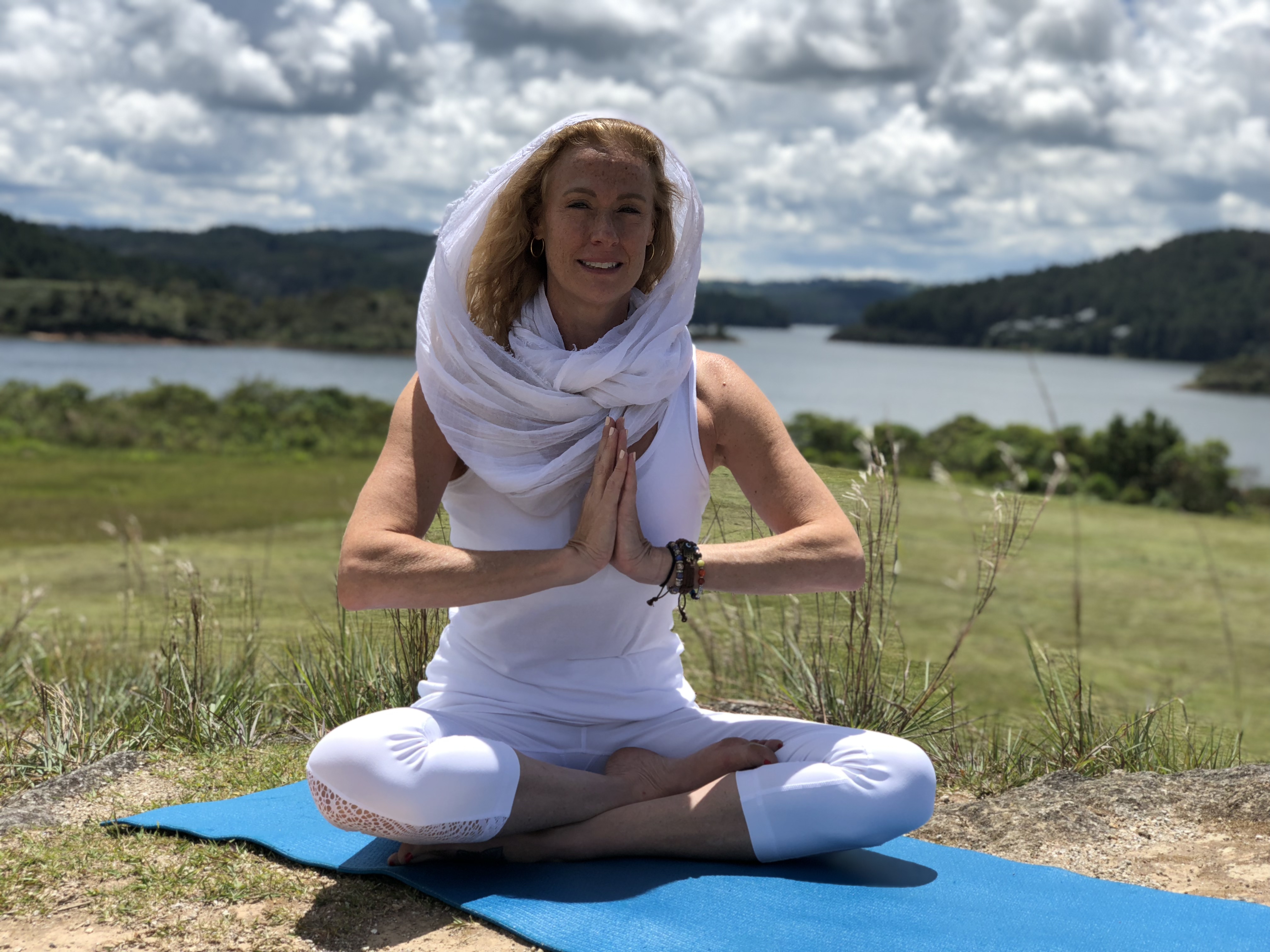 Victoria in white, seated in prayer pose outdoors by a lake